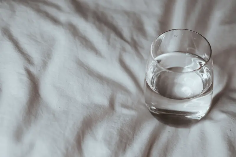 Minimalistic shot of a glass of water resting on a white duvet, taken indoors.