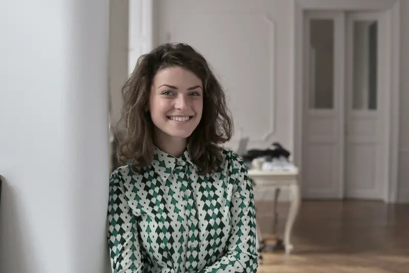 A cheerful woman with curly hair in a checkered shirt standing indoors, creating a warm and inviting atmosphere.