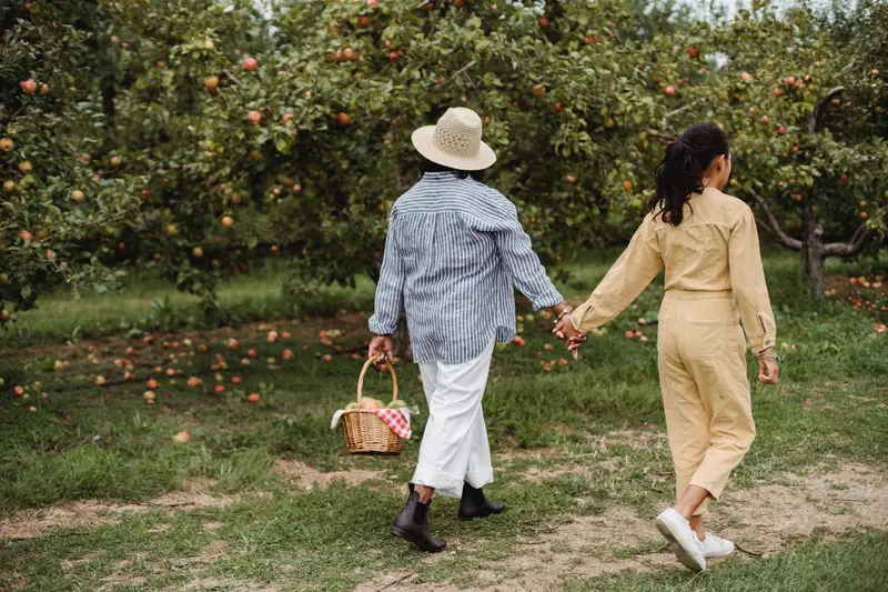 A mother and daughter walking an orchard row with a basket of apples
