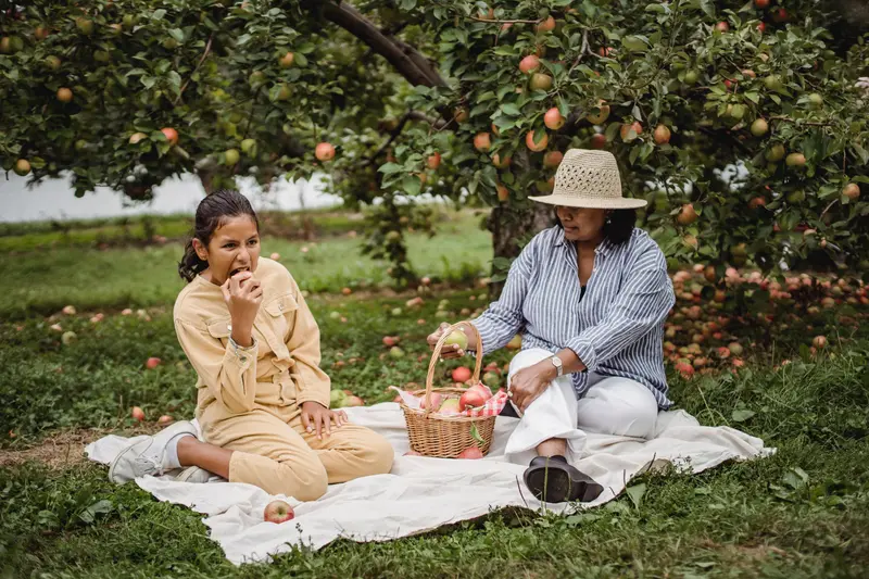 Full body of Hispanic mum sitting with basket of fresh fruits on blanket with daughter eating apple