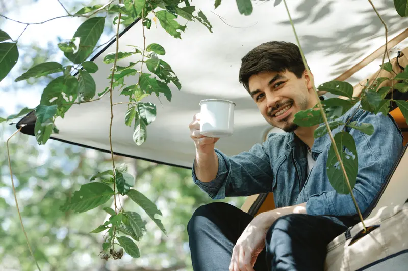 Positive young bearded male camper in casual clothes smiling and raising mug of aromatic coffee while relaxing in tent on sunny day in green forest