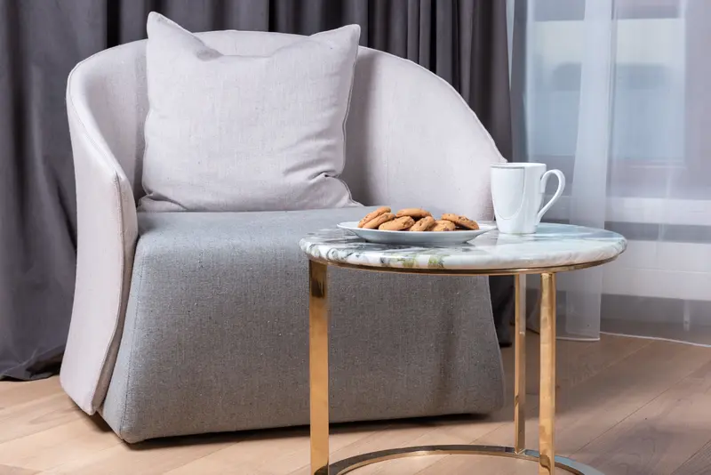 Comfortable armchair with cushion placed near window in cozy living room with plate of sweet cookies and cup of coffee arranged on marble table