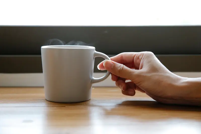 Crop anonymous female holding white cup of freshly brewed coffee placed on wooden table