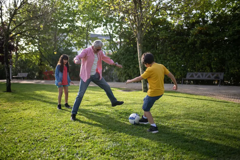 A grandfather playing soccer with his grandchildren on a sunny lawn