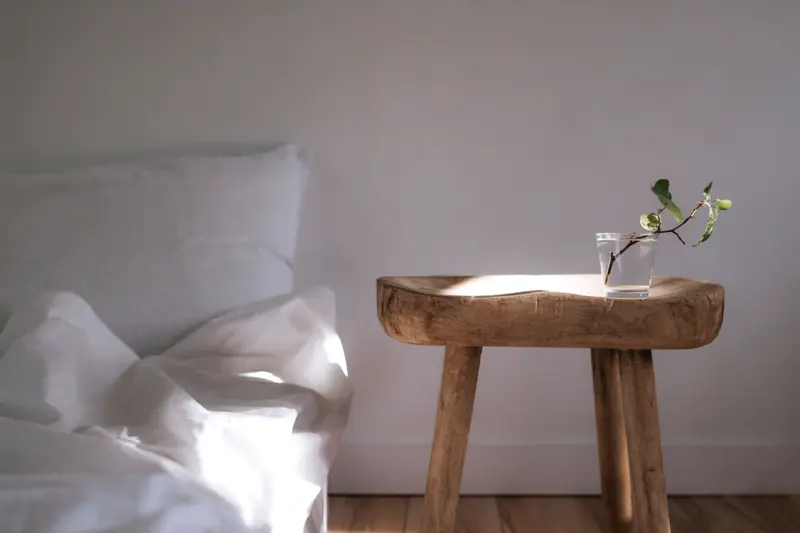 Sunlit minimalist bedroom featuring white linens and a wooden side table with a plant.