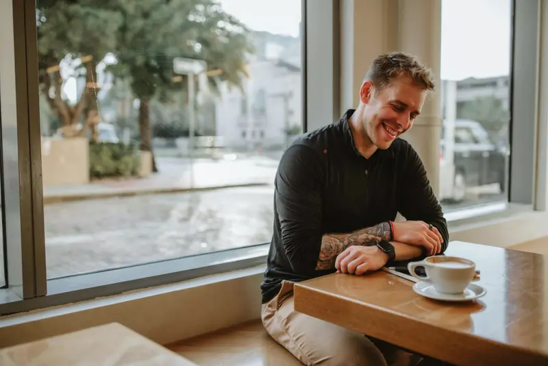 Man with tattoos smiling while enjoying coffee in a cozy cafe by the window.